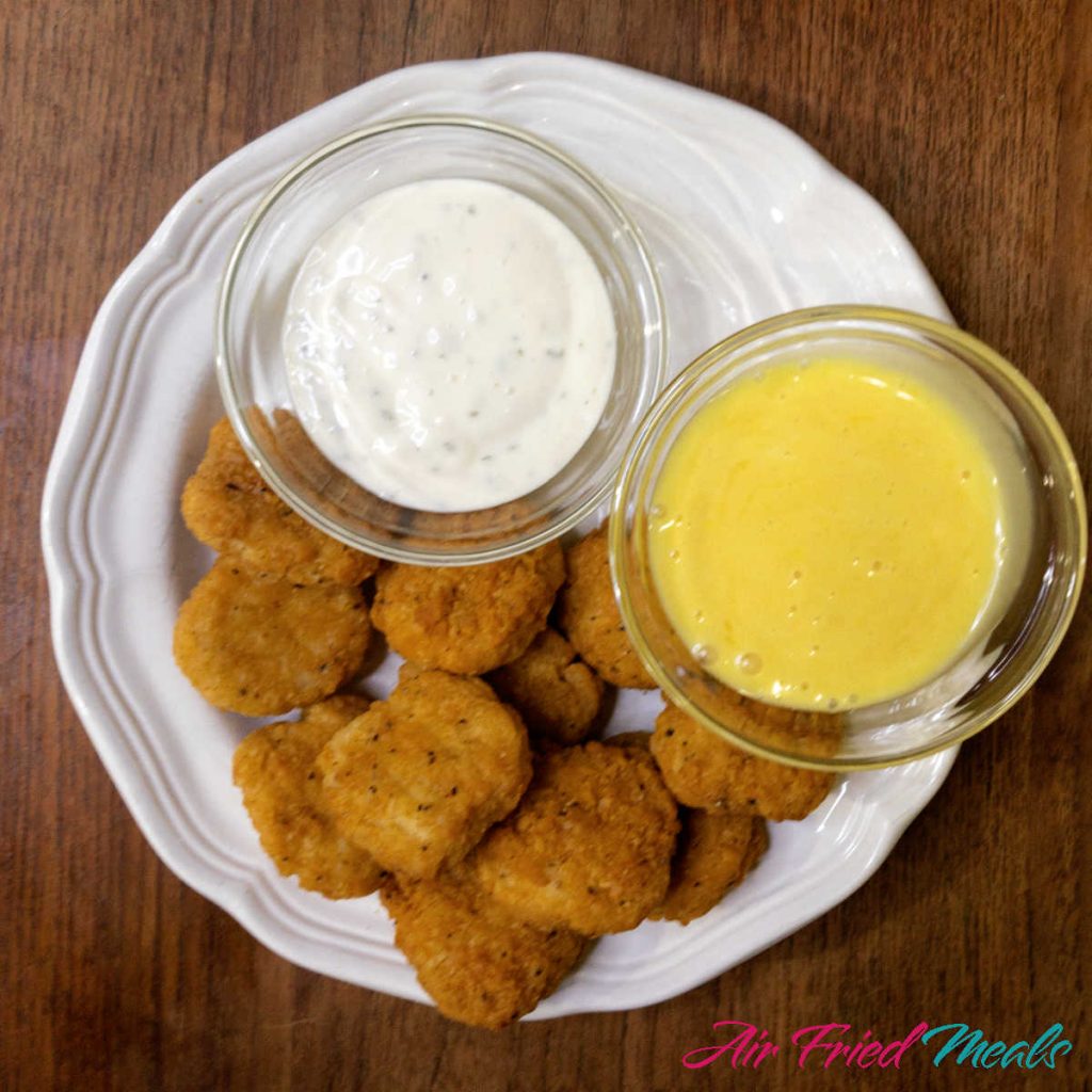 Top down view of a white plate with cooked chicken nuggets and a small bowl of ranch and a small bowl of honey mustard on plate as well.