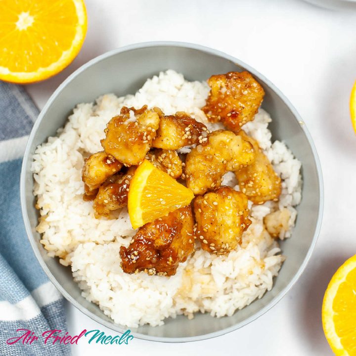 Bowl of orange chicken with rice underneath and a piece of orange on top.