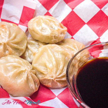 Frozen dumplings cooked in the air fryer on a red and white checkered paper with a small dipping container of soy sauce.